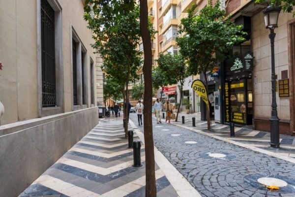 Encantadora calle urbana con pavimento, árboles y personas caminando, creando una vibrante atmósfera de verano.